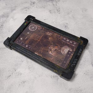 Può includere: Un tablet rettangolare grigio scuro con un display di mappa dettagliato. Il tablet ha un aspetto robusto e usurato con accenti verdi. Lo schermo mostra una mappa marrone e beige con varie icone e una bussola.