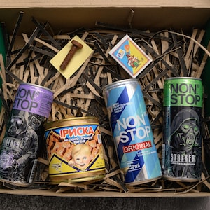 May include: A green box filled with shredded paper, containing several cans of energy drinks and a small tin of candy. The energy drink cans are purple, blue, and green, with the text "NON STOP" and graphic designs. A small bolt and a sticker are also in the box.