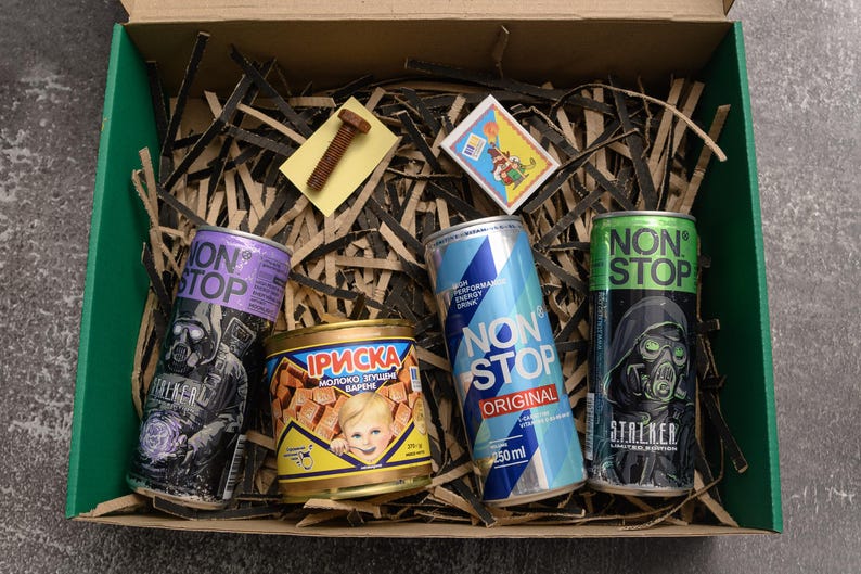 May include: A green box filled with shredded paper, containing several cans of energy drinks and a small tin of candy. The energy drink cans are purple, blue, and green, with the text "NON STOP" and graphic designs. A small bolt and a sticker are also in the box.