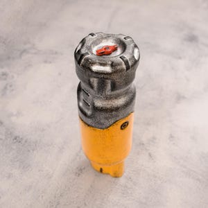 May include: A cylindrical prop featuring a weathered silver metallic top section with a small red lever and a textured yellow base. The object has a distressed finish with visible dark grey shading and a rugged, industrial appearance throughout.