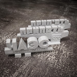 Puede incluir: Una escultura gris claro, impresa en 3D, con texto cirílico que incluye "ЧАЭС" y "ПРИПЯТЬ", junto con el año "1970". La escultura también incluye el texto "ИМЕНИ В.И. ЛЕНИНА". El diseño tiene una textura moteada.