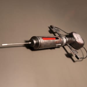 May include: A steampunk-style syringe with a metallic finish. The syringe features a long needle, a cylindrical barrel with a red liquid visible inside, and a gauge on the end. The design incorporates mechanical details and aged textures.