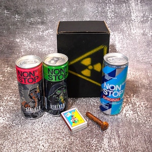 Op de afbeelding: Drie aluminium blikjes van 250 ml Non Stop energiedrank in rood, groen en blauw, een zwarte kartonnen doos met een geel stralingssymbool, een klein rechthoekig luciferdoosje met een kleurrijke afbeelding en een roestige metalen bout.