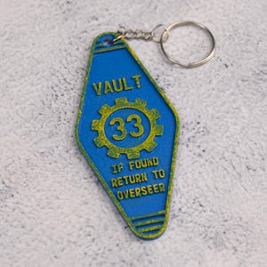 Può includere: Un portachiavi blu e giallo con la scritta "VAULT 33" e "IF FOUND RETURN TO OVERSEER". Il portachiavi ha un design a chiave da motel retrò e un portachiavi in metallo.