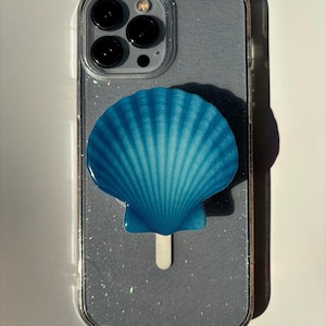 May include: A clear phone case with a glittery gray backing and a vibrant blue seashell-shaped phone grip. The phone case has a clear border and a camera cutout.
