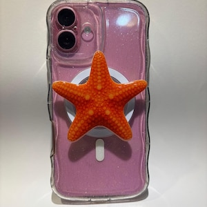 May include: A clear smartphone case with a pink glittery back panel and a dual-camera cutout. Attached to the center is a bright orange, textured starfish-shaped phone grip. The starfish features a detailed, bumpy surface and a vibrant orange color.