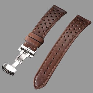 May include: Brown leather watch strap with a stainless steel clasp. The strap features a perforated design and visible stitching along the edges. The watch band is shown against a gray background.