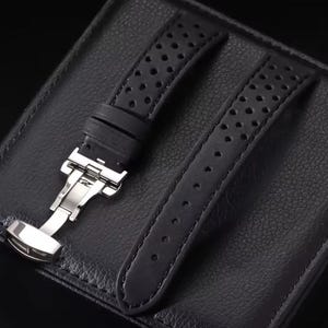 May include: Black watch strap with a perforated design. The strap features a stainless steel clasp and is displayed on a textured black surface. The strap is made of a durable material.