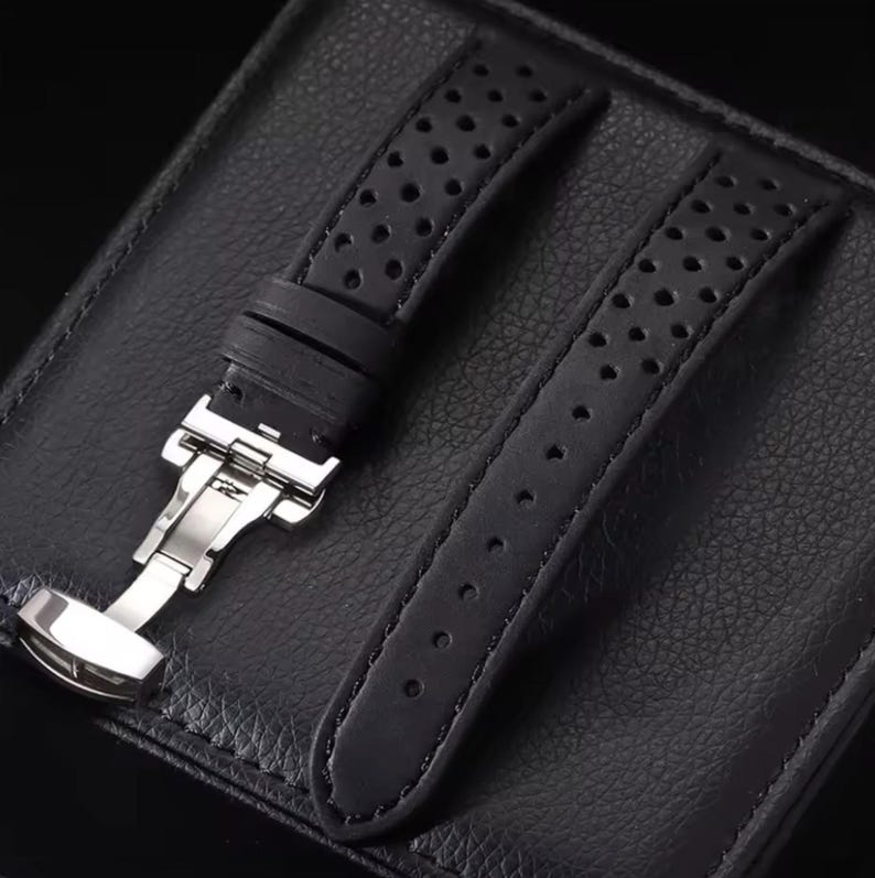 May include: Black watch strap with a perforated design. The strap features a stainless steel clasp and is displayed on a textured black surface. The strap is made of a durable material.