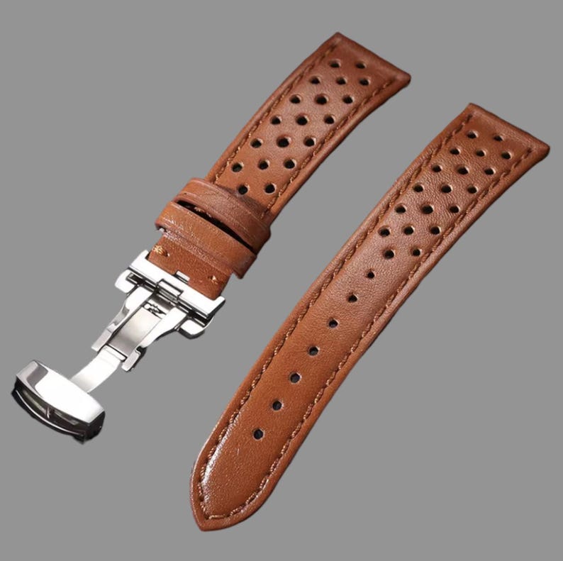 May include: Brown leather watch strap with a stainless steel clasp. The strap has a perforated design and is shown against a gray background. The watch band is new and unused.