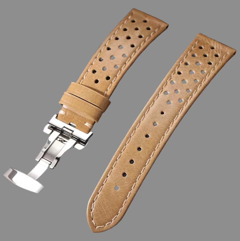 May include: Tan leather watch strap with a stainless steel clasp. The strap features a perforated design and white stitching along the edges. The watch band is shown against a gray background.