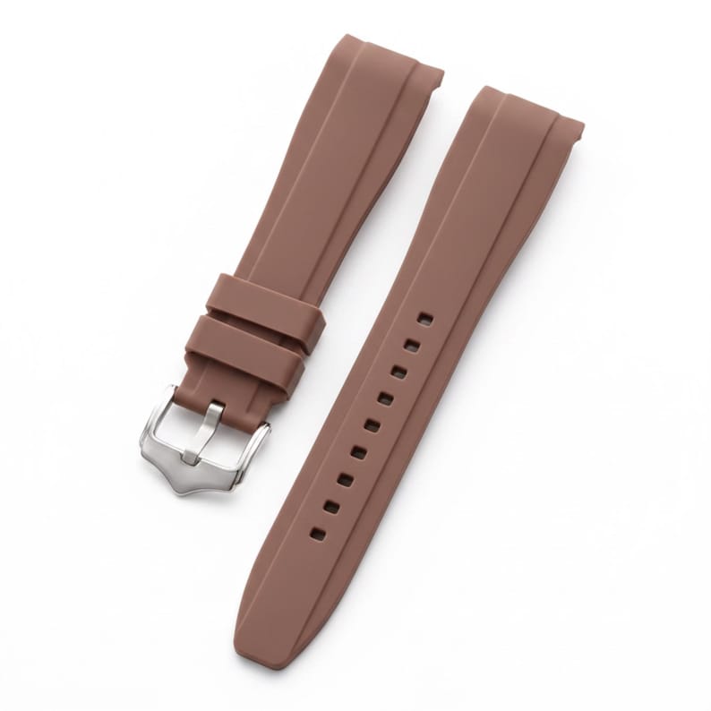 K&ouml;nnte beinhalten: Ein braunes Uhrenarmband aus Kautschuk mit glatter, matter Oberfl&auml;che, einer silberfarbenen Metallschnalle und zwei passenden braunen Kautschukschlaufen. Das Armband verf&uuml;gt &uuml;ber eine Reihe rechteckiger Einstelll&ouml;cher entlang des l&auml;ngeren Teils.