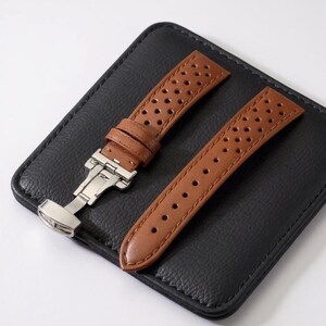 May include: A two-piece tan watch strap featuring a perforated design with small circular cutouts. The strap includes a silver-toned metal deployment clasp with a folding mechanism and matching tan stitching along the edges of the leather pieces.