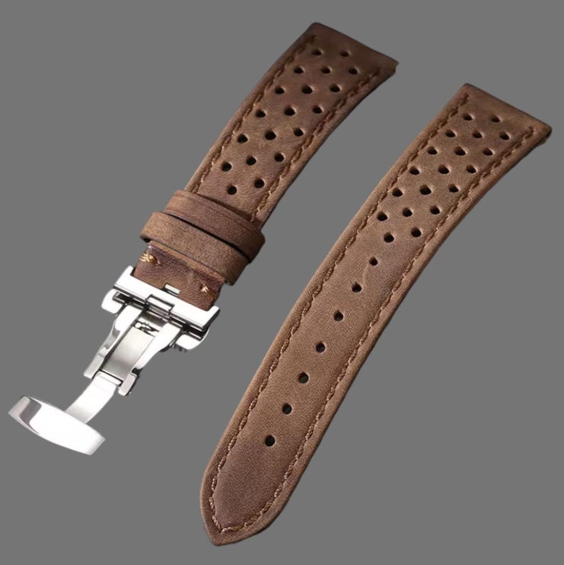 May include: Two-piece brown leather watch strap with a stainless steel clasp. The strap has a perforated design and visible stitching along the edges. The leather has a distressed appearance.
