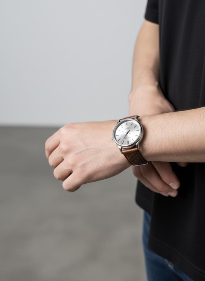 May include: A wristwatch with a round silver-toned dial, thin black hour markers, and slender hands. The watch features a brown leather strap with a perforated pattern and a silver-toned buckle, secured around a wrist with a black short-sleeved shirt.