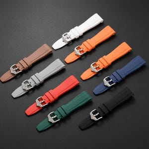K&ouml;nnte beinhalten: Eine Sammlung von neun Silikon-Uhrenarmb&auml;ndern in verschiedenen Unifarben, darunter Wei&szlig;, Orange, Braun, Grau, Rot, Dunkelgr&uuml;n, Blau und Schwarz. Jedes Armband verf&uuml;gt &uuml;ber eine silberfarbene Metallschnalle und eine passende Schlaufe.