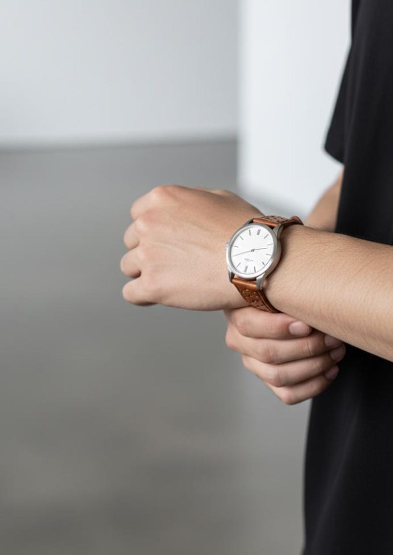 May include: A wristwatch with a round silver-tone case and a white dial featuring simple black hour markers and thin hands. The watch is attached to a tan brown leather strap that has a perforated pattern along its length and a buckle closure.