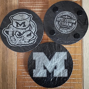 May include: Three round black slate coasters with etched designs. One coaster features a cartoonish mascot with the letter "M" inside a pot. Another has the text "Little Custom Charm". The third coaster displays a large "M".