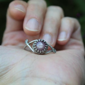 May include: A silver ring featuring a round, cabochon-cut mauve stone set in a crown-like bezel. The band is composed of two thin, curved silver wires that meet at the setting, creating an open, eye-shaped design around the central gemstone.