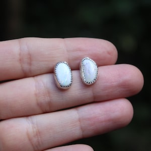 May include: A pair of oval-shaped stud earrings featuring smooth, iridescent white stones set in silver bezels with a decorative notched edge. The stones have a milky, semi-translucent appearance and are displayed against a neutral background.
