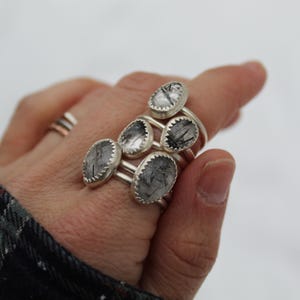 May include: A hand displays several silver rings with oval, clear stones featuring black inclusions. The rings are stacked on the fingers, showcasing the unique stone patterns. A simple silver band is also visible.