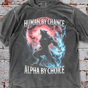 May include: Pepper-colored t-shirt featuring a werewolf graphic with the text "HUMAN BY CHANCE" above and "ALPHA BY CHOICE" below. The design includes a red moon, bats, and blue lightning. The shirt is a crew neck.