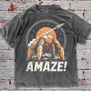 May include: Pepper-colored t-shirt with a graphic of a crab-like creature launching a rocket from a planet, with the word "AMAZE!" in white. The design features orange, white, and black elements, set against a dark gray background.