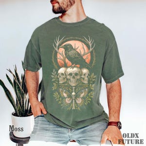 May include: Moss green t-shirt featuring a graphic design with a raven perched atop three skulls, a butterfly, and a full moon. The design includes antlers and foliage. The text "OLDX FUTURE" is printed on the bottom right.