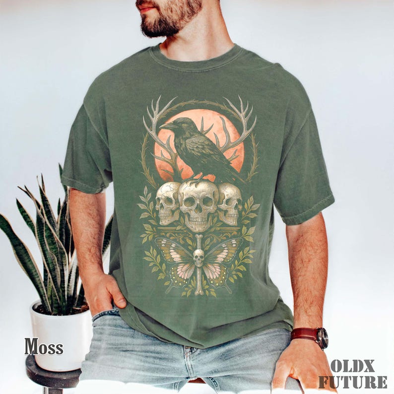 May include: Moss green t-shirt featuring a graphic design with a raven perched atop three skulls, a butterfly, and a full moon. The design includes antlers and foliage. The text "OLDX FUTURE" is printed on the bottom right.