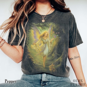 May include: Dark gray t-shirt featuring a fantasy illustration of a fairy in a forest setting. The fairy has blonde hair, a white and yellow dress, and butterfly wings. A rabbit is at the bottom of the image. The word "Pepper" is at the bottom left.
