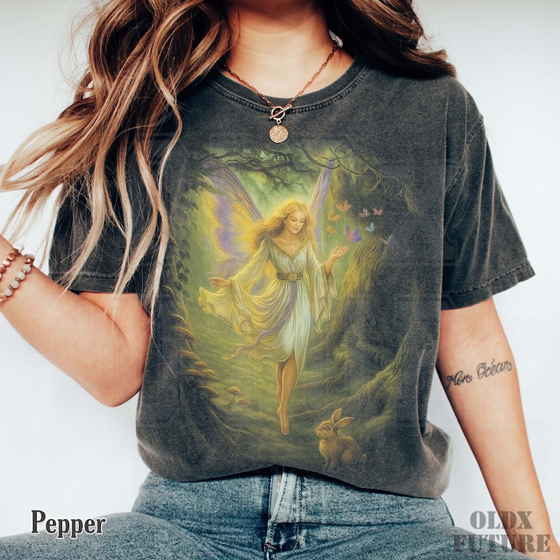 May include: Dark gray t-shirt featuring a fantasy illustration of a fairy in a forest setting. The fairy has blonde hair, a white and yellow dress, and butterfly wings. A rabbit is at the bottom of the image. The word "Pepper" is at the bottom left.