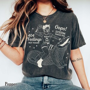 May include: Dark gray t-shirt with a graphic of a person riding an alligator, with the text "404 Feelings Not Found" and "Oops! System Error 1999". The shirt also has the words "Reboot My Soul".