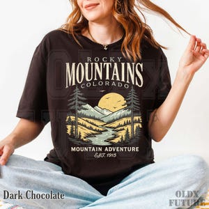 Puede incluir: Camiseta de color chocolate oscuro con un dise&ntilde;o de paisaje monta&ntilde;oso y el texto "Rocky Mountains Colorado Mountain Adventure Est. 1915". El gr&aacute;fico incluye un r&iacute;o, &aacute;rboles y un sol.