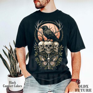 May include: Black comfort colors t-shirt featuring a detailed graphic. The design includes a raven perched atop three skulls, with a butterfly and antlers. The text "OLDX FUTURE" is visible. The shirt is a dark color.