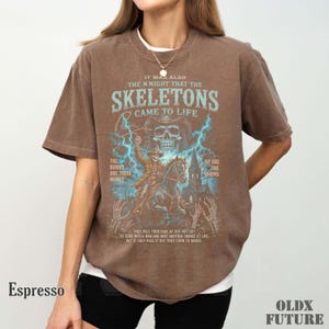 May include: A brown short-sleeve t-shirt featuring a graphic print of a skeleton cowboy on horseback with blue lightning, a skull, and text that reads: IT WAS ALSO THE NIGHT THAT THE SKELETONS CAME TO LIFE, THE BONES ARE THEIR MONEY, SO ARE THE WORMS.