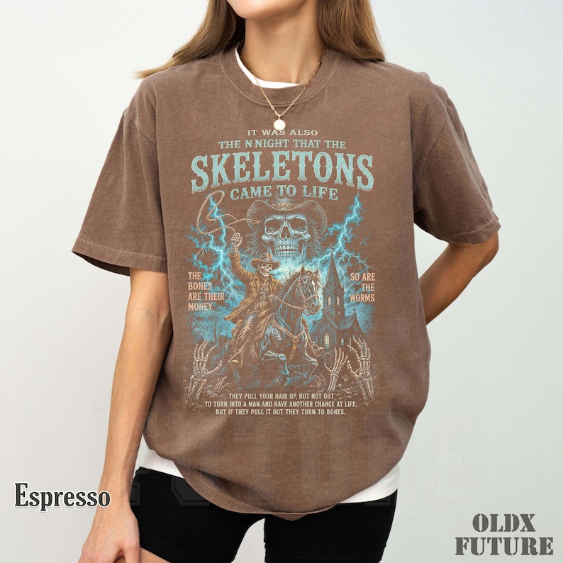 May include: A brown short-sleeve t-shirt featuring a graphic print of a skeleton cowboy on horseback with blue lightning, a skull, and text that reads: IT WAS ALSO THE NIGHT THAT THE SKELETONS CAME TO LIFE, THE BONES ARE THEIR MONEY, SO ARE THE WORMS.