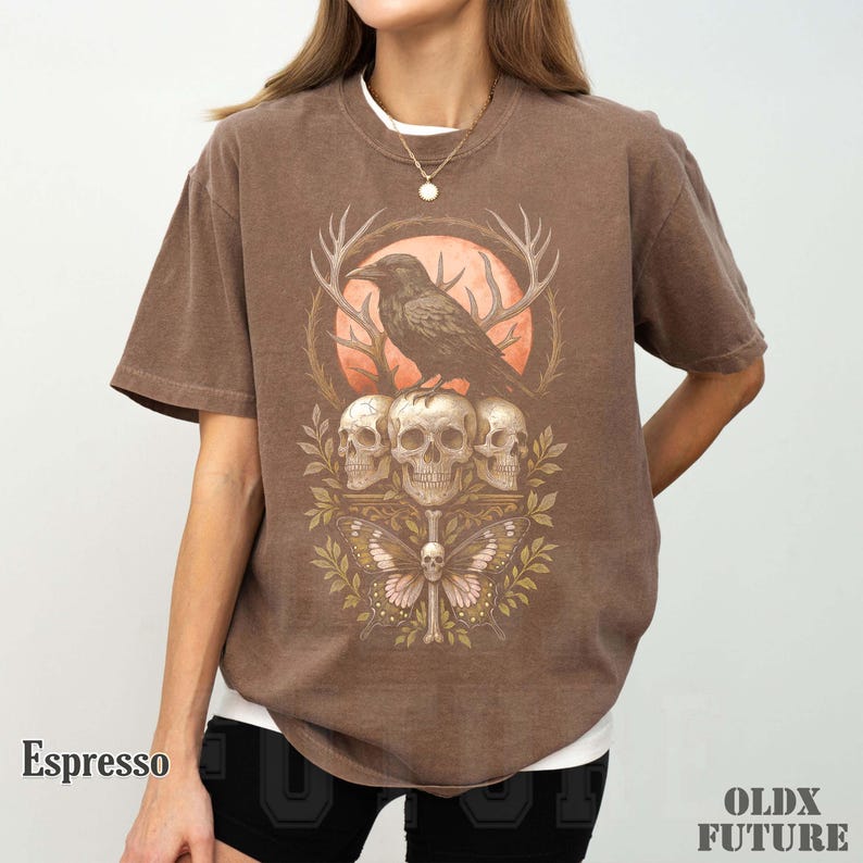 May include: Espresso-colored t-shirt featuring a graphic design of a raven perched atop three skulls, with a butterfly and antlers. The shirt has short sleeves and a crew neck. The text "OLDX FUTURE" is in the bottom right corner.