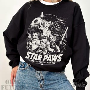 May include: A black long-sleeve sweatshirt featuring a white graphic print of cats dressed as space characters. The design includes the text STAR PAWS and MAY THE PURR BE WITH YOU, along with a space station, a starfighter, and various cat characters.