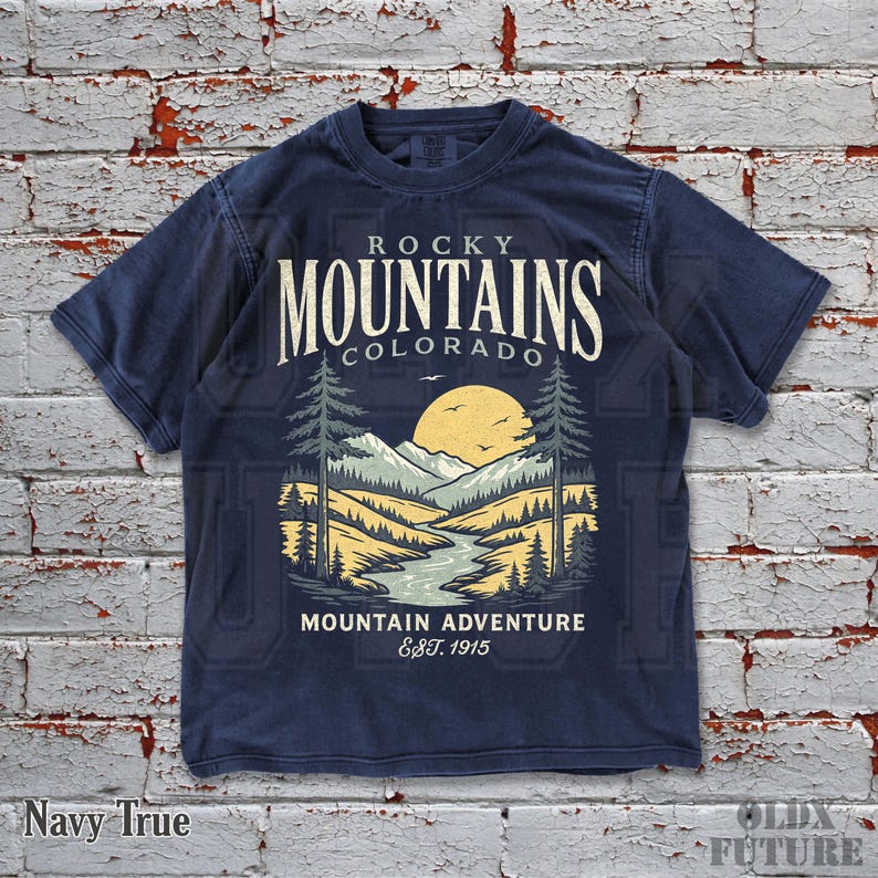 Puede incluir: Camiseta azul marino con un dise&ntilde;o de paisaje monta&ntilde;oso, un r&iacute;o y un sol poniente. El texto "Rocky Mountains Colorado" y "Mountain Adventure Est. 1915" est&aacute; impreso. La camiseta est&aacute; etiquetada como "Navy True".
