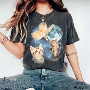 May include: Dark gray t-shirt featuring a graphic of three kittens in space with a blue moon. The kittens are various shades of brown and tan. The word "Pepper" is printed on the bottom left of the shirt.