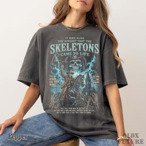May include: A charcoal gray short-sleeve t-shirt featuring a graphic print of a skeleton cowboy riding a horse in front of a church. The text reads: IT WAS ALSO THE N NIGHT THAT THE SKELETONS CAME TO LIFE, THE BONES ARE THEIR MONEY, SO ARE THE WORMS.