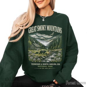 May include: A forest green crewneck sweatshirt featuring a graphic print of the Great Smoky Mountains. The design includes a cabin, a stream, bears, and an eagle, with text reading Great Smoky Mountains, Tennessee & North Carolina, USA, National Park, Est. 1934.