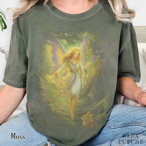 May include: Moss green t-shirt featuring a fantasy illustration of a fairy in a forest setting. The fairy has long blonde hair, a white dress, and butterfly wings. Butterflies and a rabbit are also depicted in the artwork. The word "Moss" is printed at the bottom left.