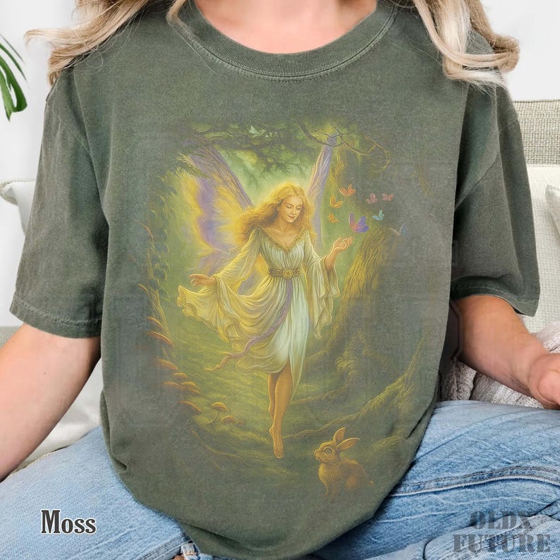 May include: Moss green t-shirt featuring a fantasy illustration of a fairy in a forest setting. The fairy has long blonde hair, a white dress, and butterfly wings. Butterflies and a rabbit are also depicted in the artwork. The word "Moss" is printed at the bottom left.