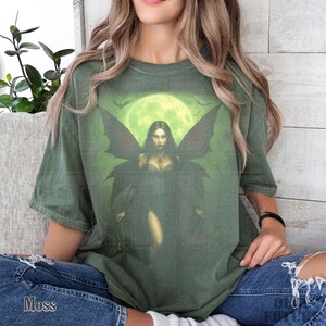 May include: Moss green t-shirt featuring a graphic of a dark fairy with large wings, set against a green moonlit background with bats. The shirt has a relaxed fit and is made of a soft material. The word "Moss" is printed on the bottom left.