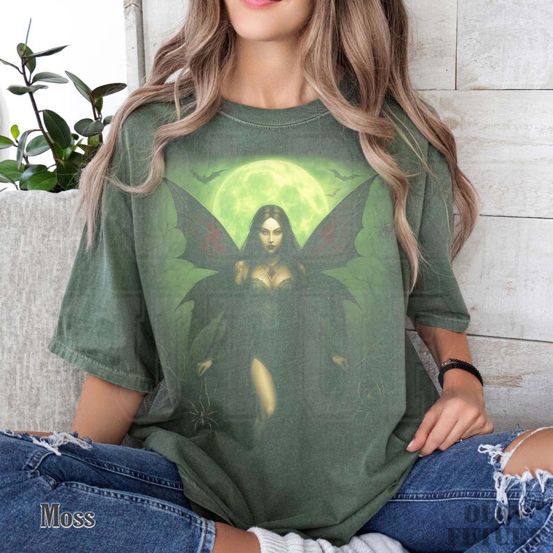 May include: Moss green t-shirt featuring a graphic of a dark fairy with large wings, set against a green moonlit background with bats. The shirt has a relaxed fit and is made of a soft material. The word "Moss" is printed on the bottom left.