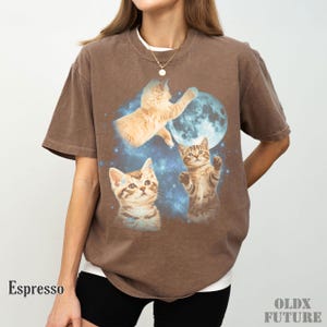 May include: Espresso-colored t-shirt featuring a whimsical design of three kittens reaching for a blue moon in a starry sky. The shirt has a relaxed fit and the word "Espresso" is printed on the bottom left.