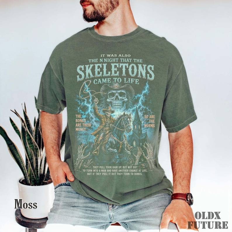 May include: An olive green short-sleeve t-shirt featuring a graphic print of a skeleton cowboy riding a horse with lightning, a church, and the text: IT WAS ALSO THE NIGHT THAT THE SKELETONS CAME TO LIFE. THE BONES ARE THEIR MONEY SO ARE THE WORMS.