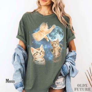 May include: Moss green t-shirt featuring a graphic of three kittens, a full moon, and a starry background. The shirt has rolled-up sleeves and the word "Moss" is printed on the lower left. The brand name "OLDX FUTURE" is printed on the lower right.