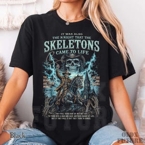 May include: A black t-shirt featuring a graphic print of a skeleton cowboy riding a horse in front of a church. The text reads: IT WAS ALSO THE N NIGHT THAT THE SKELETONS CAME TO LIFE, THE BONES ARE THEIR MONEY, SO ARE THE WORMS.